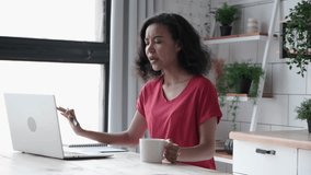 Young African American woman has video call using laptop for freelance work at kitchen. Curly-haired female in red T-shirt makes notes in paper notebook at wooden table 