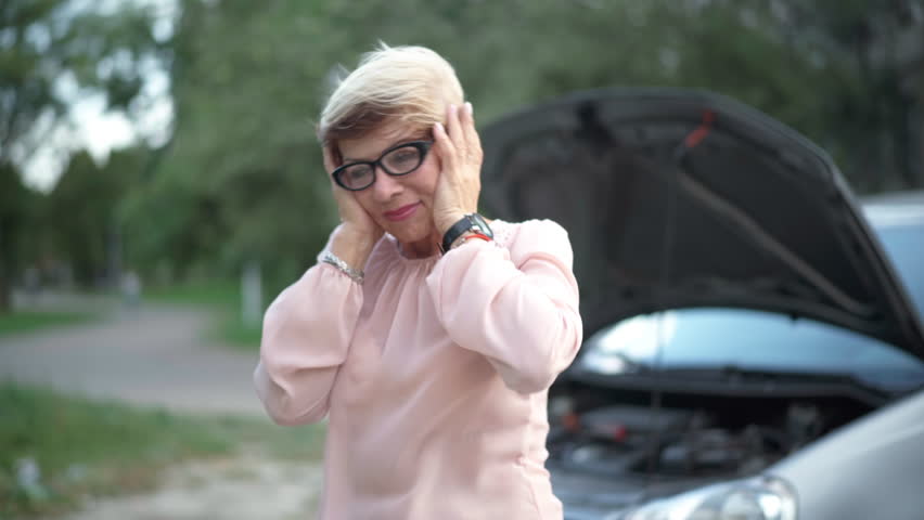 Worried stressed senior woman in eyeglasses holding head in hands standing at broken vehicle outdoors. Medium shot portrait of Caucasian retiree with broken car thinking