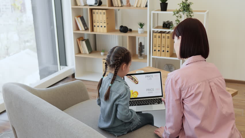 Dark haired woman and little girl using portable laptop for ordering lunch in online shop. Happy mother with lovely daughter purchasing food with discount for saving and free time at home.