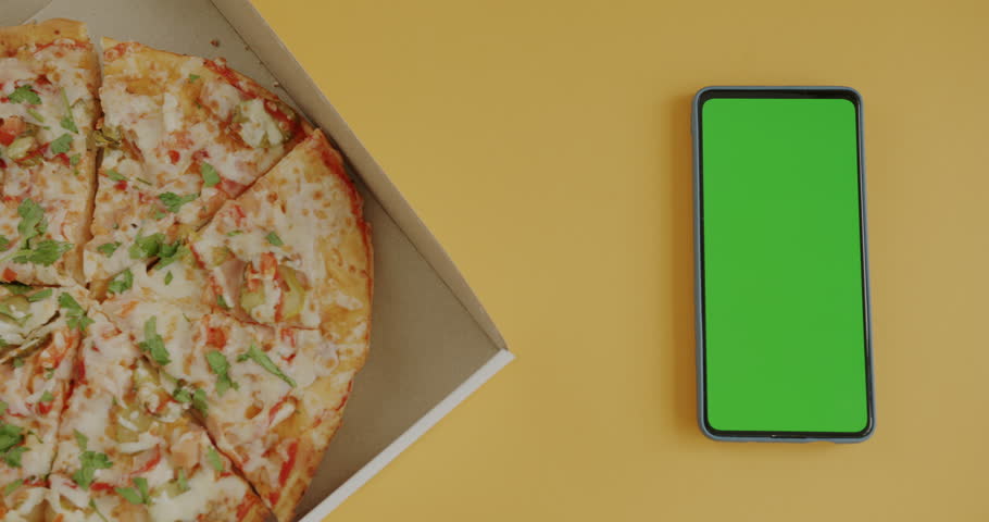 Top view of hand taking slice of pizza from box next to green screen chroma key smartphone. Food delivery and modern technology concept.