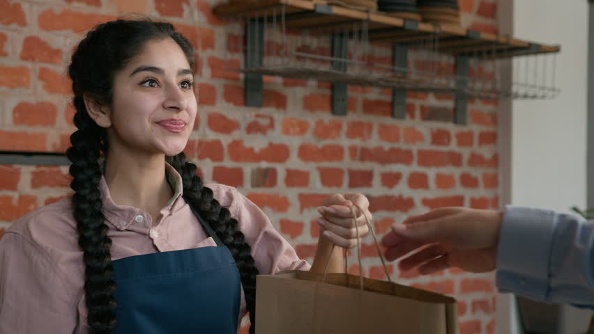 Friendly young indian woman waitress barista giving takeaway food bag to customer female seller cafe employee in apron holding takeout order delivery in cafeteria restaurant coffee shop serves client Royalty-Free Stock Footage #1101358287