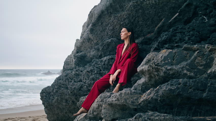 Relaxed stylish woman sitting under rock ledge in elegant red suit. Thoughtful young girl looking on gloomy dramatic ocean view. Fashionable pensive lady posing in front stone cliff at autumn seashore