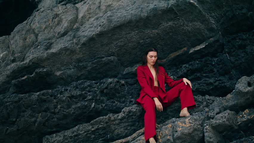 Confident beautiful woman sitting on mountain ledge wearing red provocative suit. Gorgeous sexy model posing at stone rock outdoors cloudy weekend. Serious cheeky girl looking camera on nature.