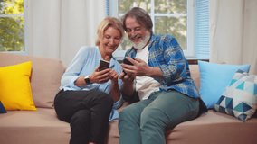 Happy senior couple use smart phone having fun with mobile app. Cheerful old retired grandparents laugh look at cellphone screen relax sit on sofa at home. Aged couple use smartphone. Realtime