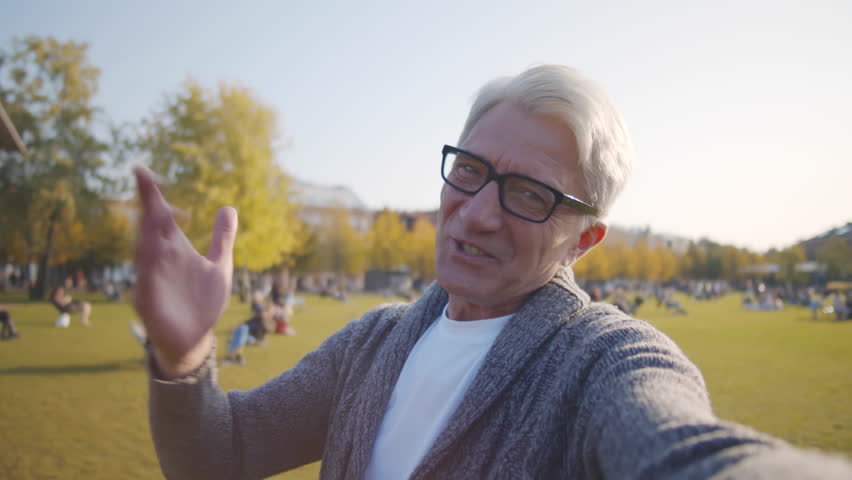 Pov shot of happy senior man talk on video chat outdoors. Mature male blogger make video talking at camera in park. Aged man have video conference in city. Realtime