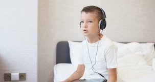 A kid boy in white T-shirt using tablet computer with headphones, siting on bed