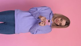 Cheerful woman using smartphone on pink background in studio. Vertical video.