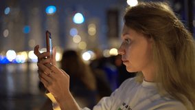 Young blonde girl stands on the embankment and shoots a video on the phone and taking selfie among the skyscrapers at night and smiling