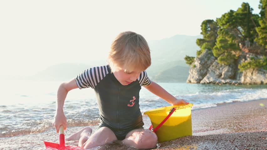 Happy child playing with sand and stones sitting on the seashore. Joyful boy have fun during a beach holiday by the clear warm sea. Leisure for kids and the whole family during school holidays Royalty-Free Stock Footage #1101381477