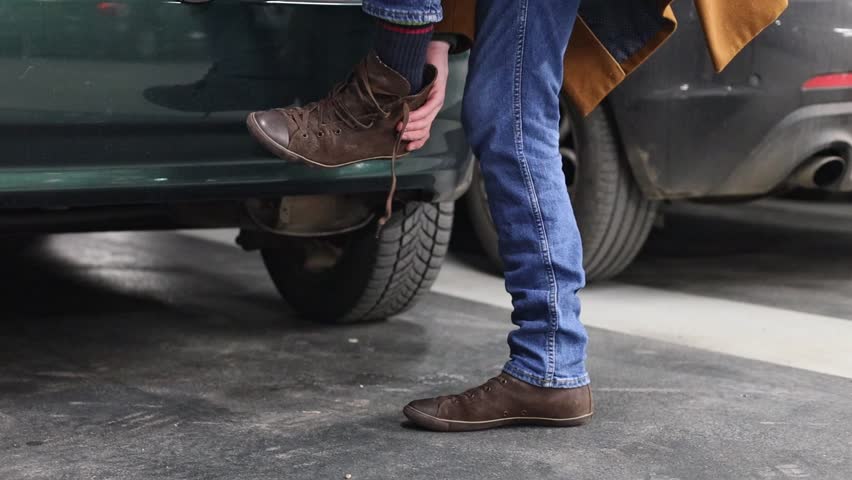 Young caucasian man in a brown coat takes off his old shoe and puts on a new one while squatting in the underground parking of a store, close-up side view. Men lifestyle concept.