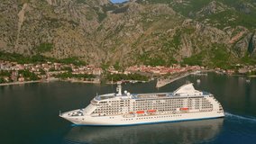 The drone captures a stunning aerial view of a white cruise ship leaving the Boka-Kotor bay in Montenegro during sunset. The calm sea and surrounding mountains create a serene atmosphere, making it a