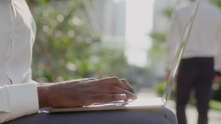 Tilt up shot of African American businessman sitting outside on sunny day, working on laptop and celebrating success with clenched fists