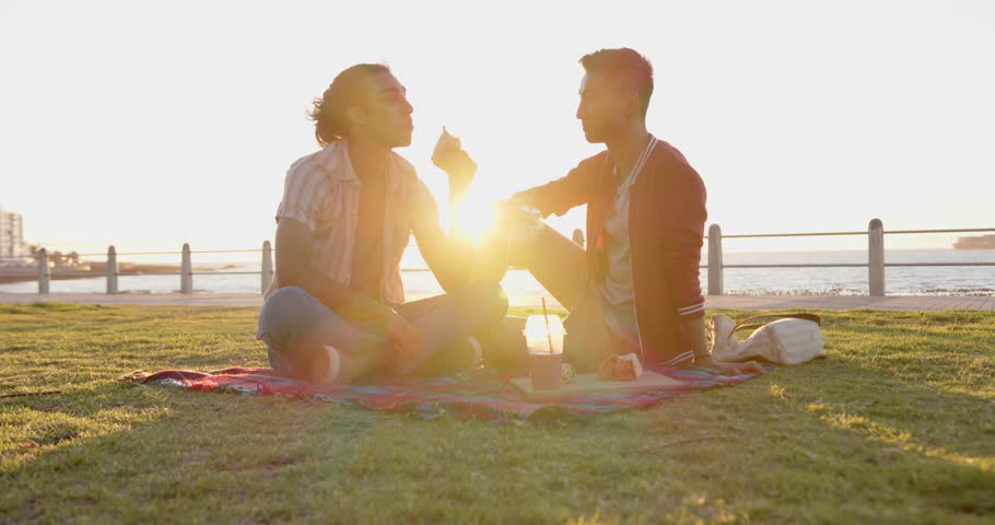 Happy diverse gay male couple having picnic at promenade by the sea, slow motion. Spending quality time, lifestyle, summer and vacation.