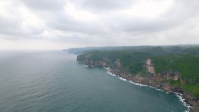aerial view of a coastline that has cliffs with dense forest in cloudy sky. Sea waves crashing the rocks at the base of the cliff make white foam- Dronie shot