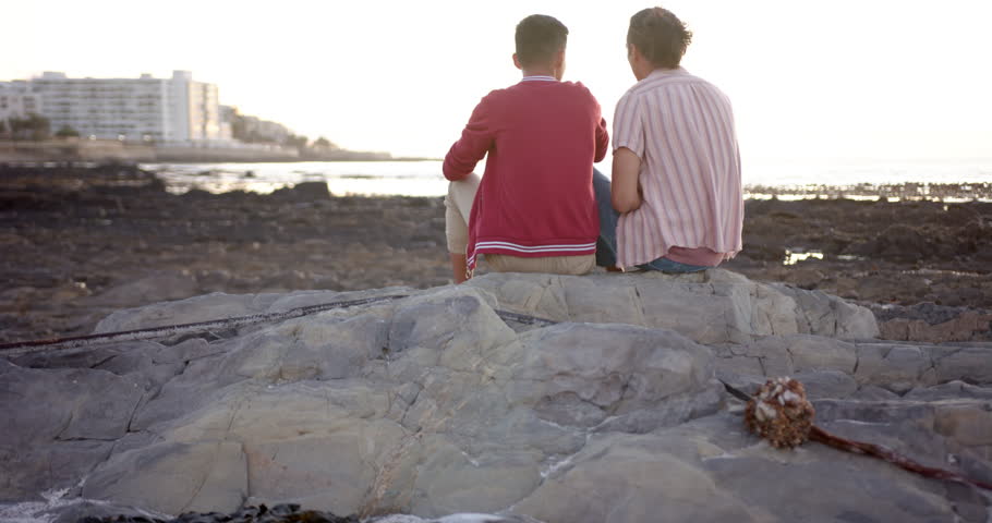 Happy diverse gay male couple sitting on rocks and embracing at beach, slow motion. Spending quality time, lifestyle, summer and vacation.