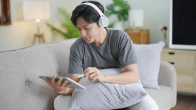 Handsome Asian man wearing headphones listening to music and using tablet sitting on sofa in holiday home