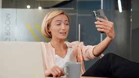Cheerful blonde office worker engages in a video call on her mobile phone, emphasizing the importance of remote communication and teamwork in the digital age. Business and technology related projects.