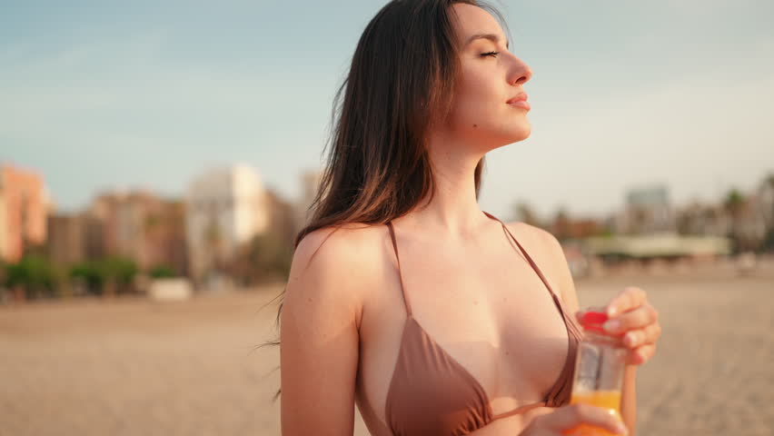 Slim beautiful brown-haired woman with long hair drinks juice from plastic bottle while standing on the beach. Gorgeous girl with sporty figure is testing the juice. slow motion