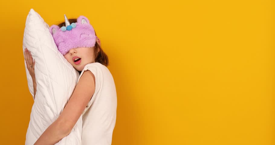 Nice teenage girl in white pyjamas with a violet sleeping mask embraces a pillow in the studio on a yellow background, ready to sleep Royalty-Free Stock Footage #1101398793