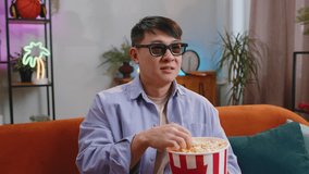 Excited asian man in 3D glasses sits on couch eating popcorn snacks and watching interesting TV serial sport game, film online social media movie content at home apartment. Guy enjoying entertainment