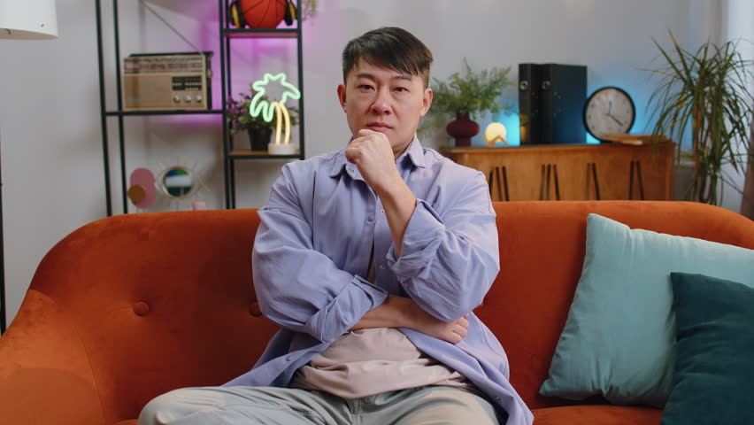 I dont know what to say. Confused asian handsome man feeling embarrassed about ambiguous question, having doubts, no idea, being clueless and uncertain at home. Chinese guy sits on sofa in living room Royalty-Free Stock Footage #1101403685