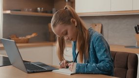Cute primary Caucasian school girl studying at kitchen with laptop. Child daughter kid schoolgirl pupil doing homework writing exercise distance learning. Children home education e-learning quarantine