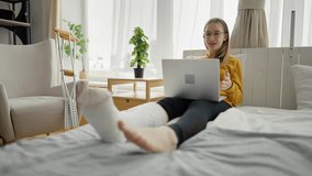 Teenage girl with broken leg in cast sits at home and use laptop to online video call conversation with friends. Young woman sitting on bed in living room next to crutches. Injury, home treatment.