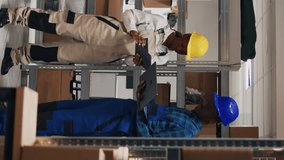 Vertical video: African american people analyzing goods on laptop and tablet, using devices to create stock inventory in storehouse space. Young man and woman working with cardboard packages and cargo