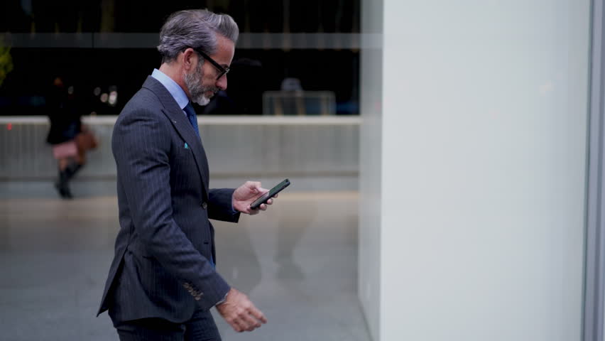 Successful mature businessman reads text message on mobile phone while walking near office building.Adult prosperous entrepreneur in suit using smartphone application for sending email and communicate Royalty-Free Stock Footage #1101412145