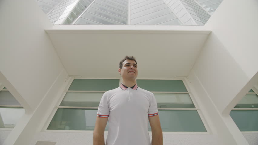 Low angle shot of teenager in white t-shirt looking serious and concerned in whitish interior. Portrait of young, handsome man feeling successful and attractive in sunglasses. High quality 4k footage