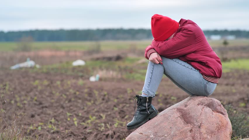 A sad teenage girl sits on a rock in a field and cries.