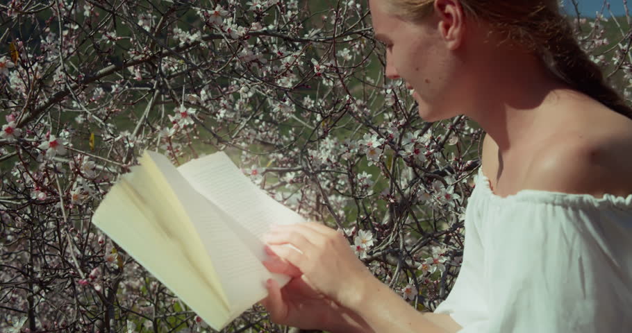 Girl reading a book outdoors. Close-up of woman face with open dairy in hands. Spring flowers blurred background.