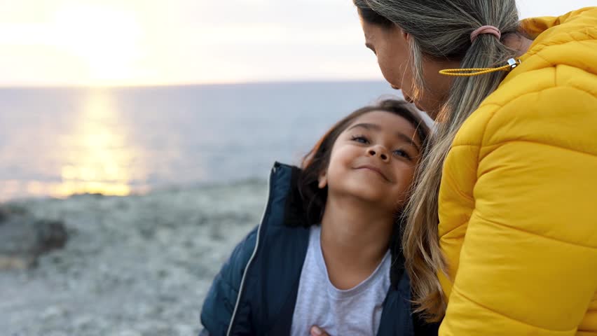 Latin mother and daughter having tender moment together on the beach at sunset during winter time