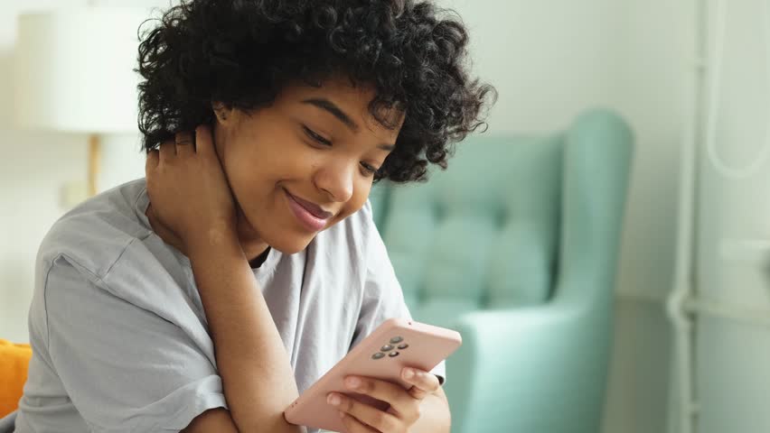 African girl holding smartphone touch screen typing scroll page at home. Woman with cell phone surfing internet using social media apps playing game. Shopping online Internet news cellphone addiction