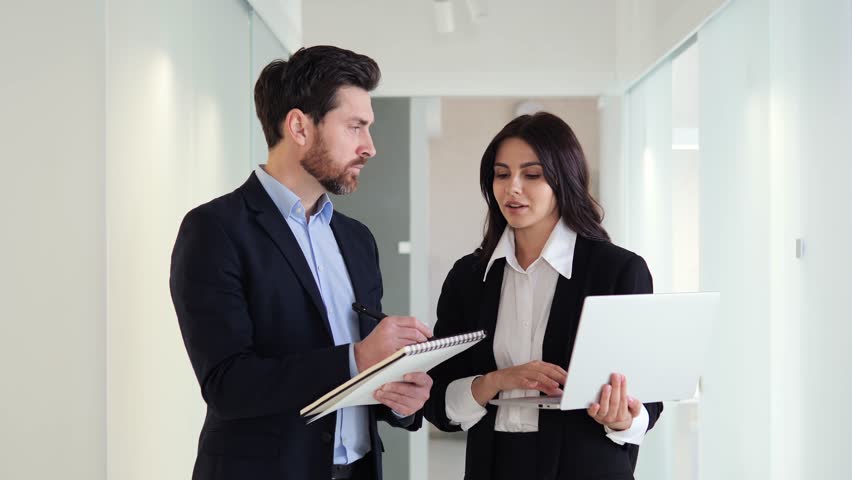 Office female male workers collaborate in the hallway, using laptops and paper documents to discuss and strategize about a project. They are determined to meet their deadline and achieve their goals.