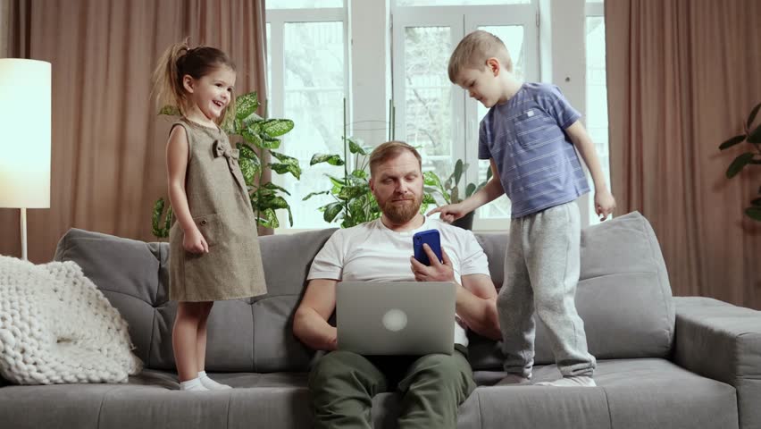 Businessman, father sitting on sofa in living room, working and talking on phone. Little boy and girl, kids jumping around and disturbing him. Concept of fatherhood, childhood, freelance, remote work Royalty-Free Stock Footage #1101435443