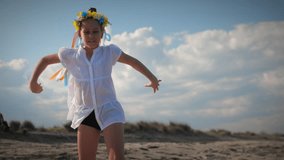 Ukrainian girl in shirt with floral wreath with multi-colored ribbons dances on coast in sunbeams