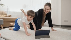 Smiling boy with mother watching online fitness lesson on tablet and doing sports exercises at home. Family healthcare, active lifestyle, parenting and child development.