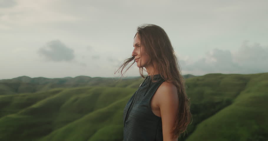 Woman enjoy green hills landscape at sunset. Long hair brunette girl looking on beautiful nature scene, hair blowing in wind. Freedom, happiness lifestyle. Travel, tourism, holiday. Slow motion 4k