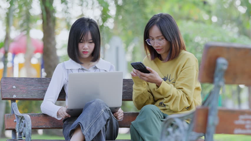Portrait of two Asian women sitting at office park while using digital smartphone and laptop.