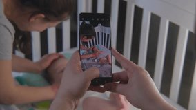 Father takes a picture or video of happy mother with newborn adorable daughter or son on mobile phone. A little baby in bodysuit lies in child crib. Concept of childhood, parenthood, love and family.