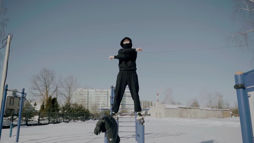 A guy in a tracksuit is standing on a horizontal bar in winter warming up. He has a hood on his head