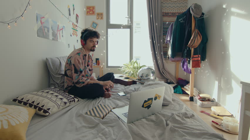 Young man in headphones listening to music on laptop, dancing and vibing in bedroom with diy decor and disco ball. Zoom Shot