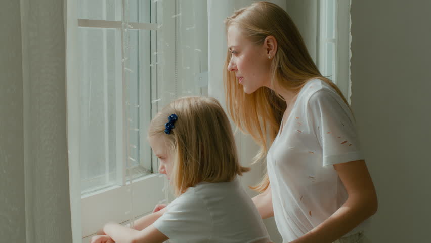 Caucasian mother with little preschool daughter standing in morning light looking window. Child kid girl pointing with finger direction to outside peeking someone mum talking with kid at home room Royalty-Free Stock Footage #1101471921