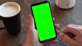 Man Use of Mobile Phone with Blank Green Screen in a Cafe on Rustic Vintage Wooden Table. Male Holding Smart Phone Empty ChromaKey Screen Mock up in Coffee Shop. Morning. Showing Mock up Video Footage