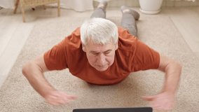 Mature senior man doing fitness exercises, gymnastics at home. Online lesson with laptop pc. Talking trainer. Recreation, well being. Elderly male exercising training, stretching. Old man working out