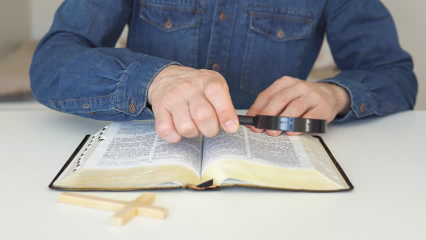 Christian catholic man with magnifying glass searching for answers in old Holy Bible. Loupe on a Bible.