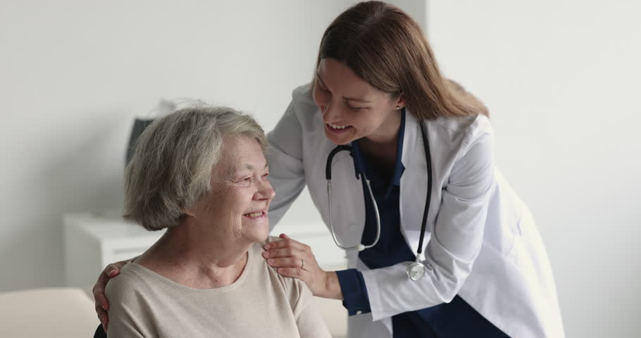 Empathetic geriatric doctor calming down positive smiling senior patient, touching shoulders, hugging, talking to elderly woman in physician office, speaking, explaining treatment details | Shutterstock HD Video #1101476269