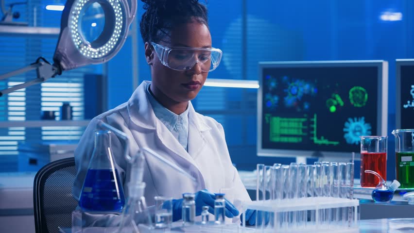 A young African American woman using a plastic pipette pipettes red and green liquid into glass test tubes and examines samples. A black female doctor works in a biochemical laboratory with blue light Royalty-Free Stock Footage #1101476675