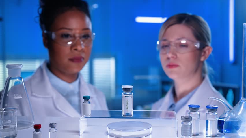 An African American female doctor and a white female laboratory assistant examine a glass vaccine vial. Medical workers in goggles and white gowns. Laboratory or hospital with blue light. Close up. Royalty-Free Stock Footage #1101476691